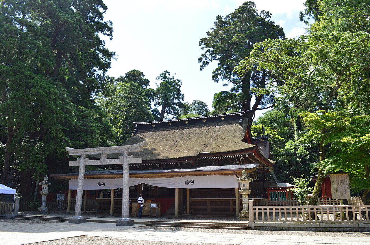 The bark-roofed Haiden worship hall of Kashima Jingu behind a small stone torii, surrounded by tall cedars
