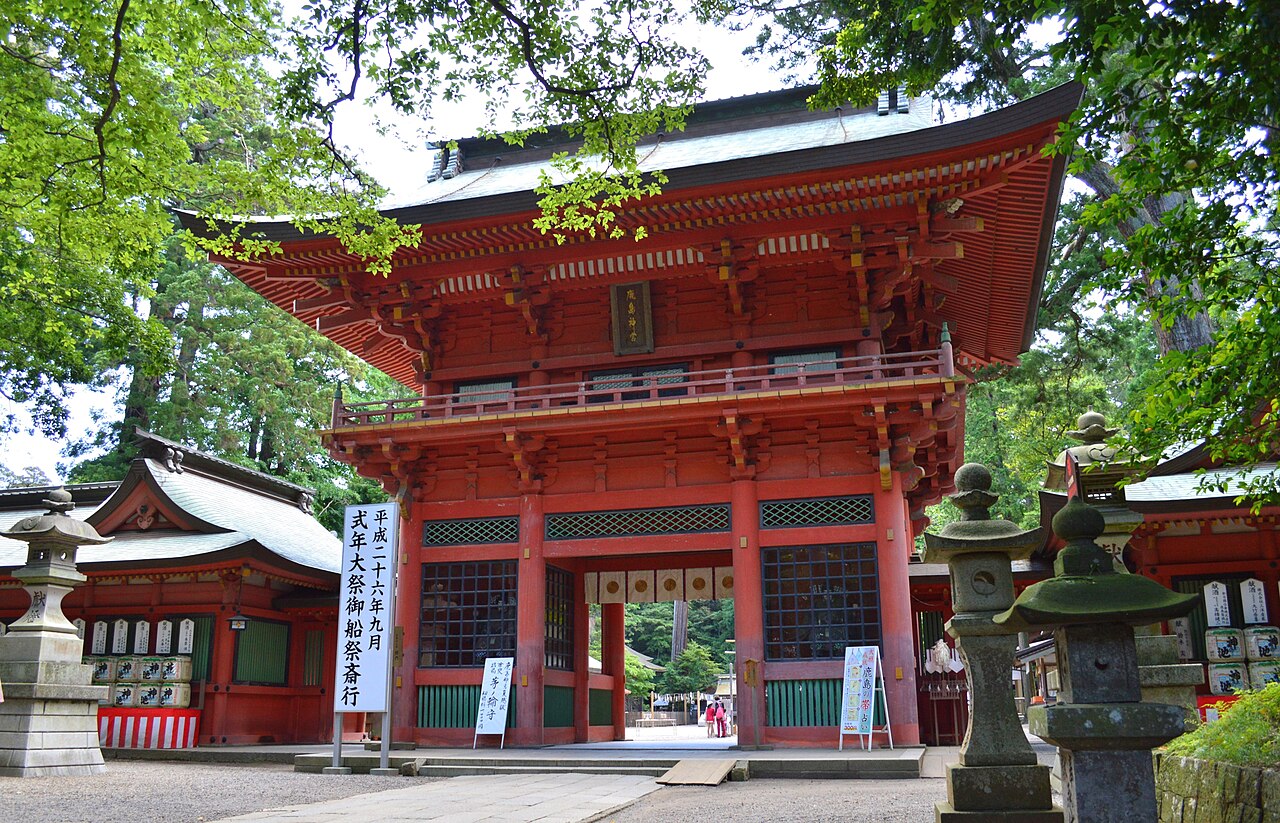 The red two-storey Romon gate of Kashima Jingu seen from the gravel courtyard, with stone lanterns and sake barrels beside it