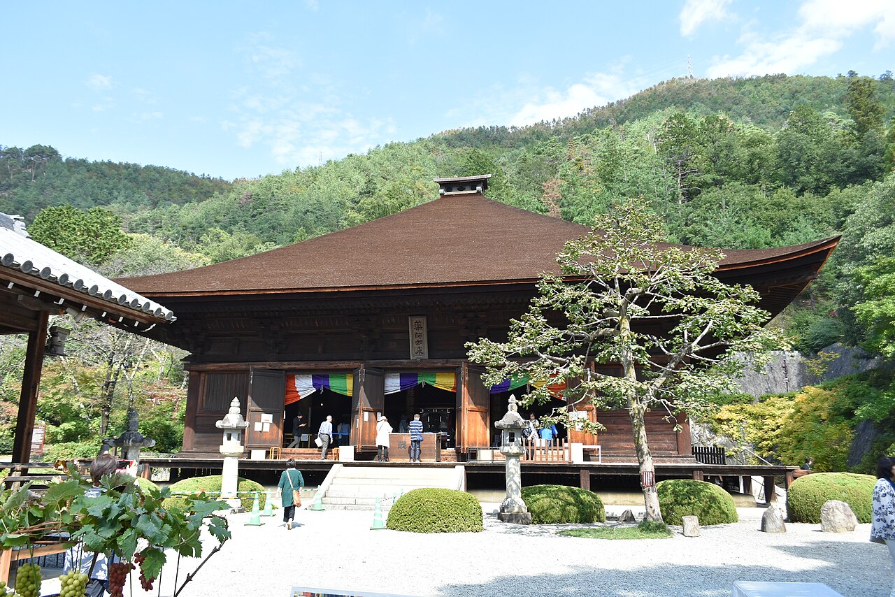The dark wooden main hall of Daizenji temple with a sweeping hipped roof, colorful hanging cloths at the front and green hills behind.