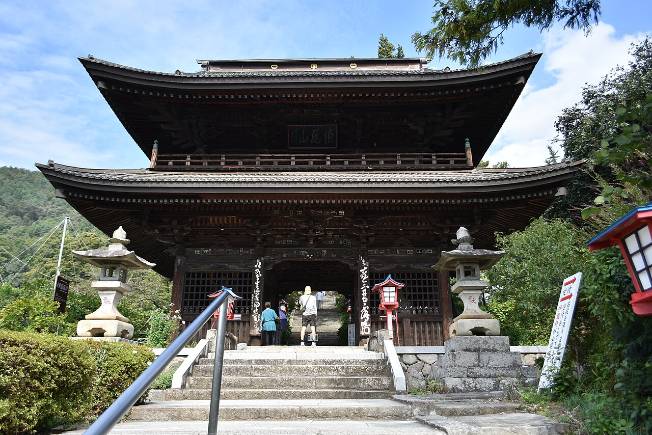 The two-story wooden gate of Daizenji temple at the top of a stone stairway, flanked by stone lanterns and red lanterns.
