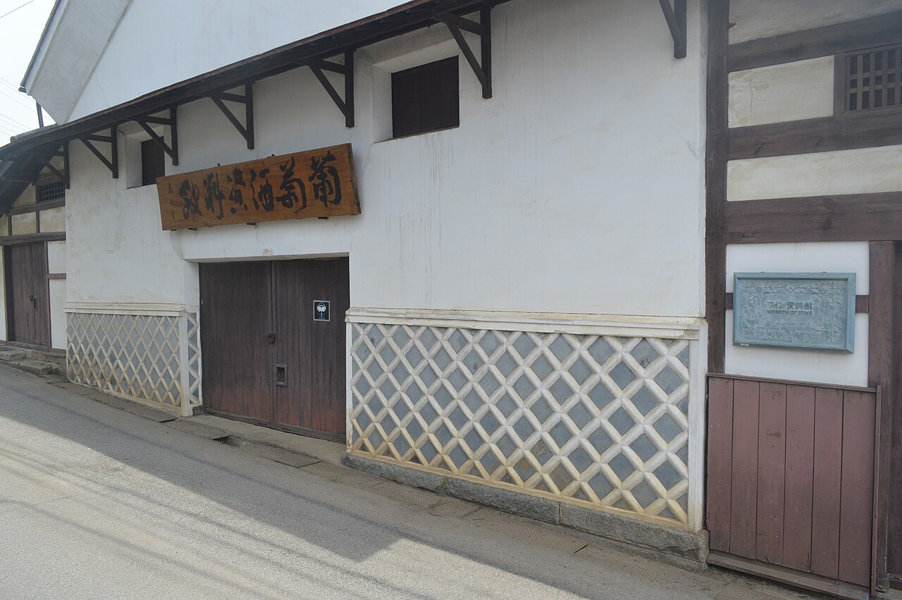 A white plaster storehouse with a carved wooden signboard and lattice-patterned lower walls at an old winery building in Katsunuma.