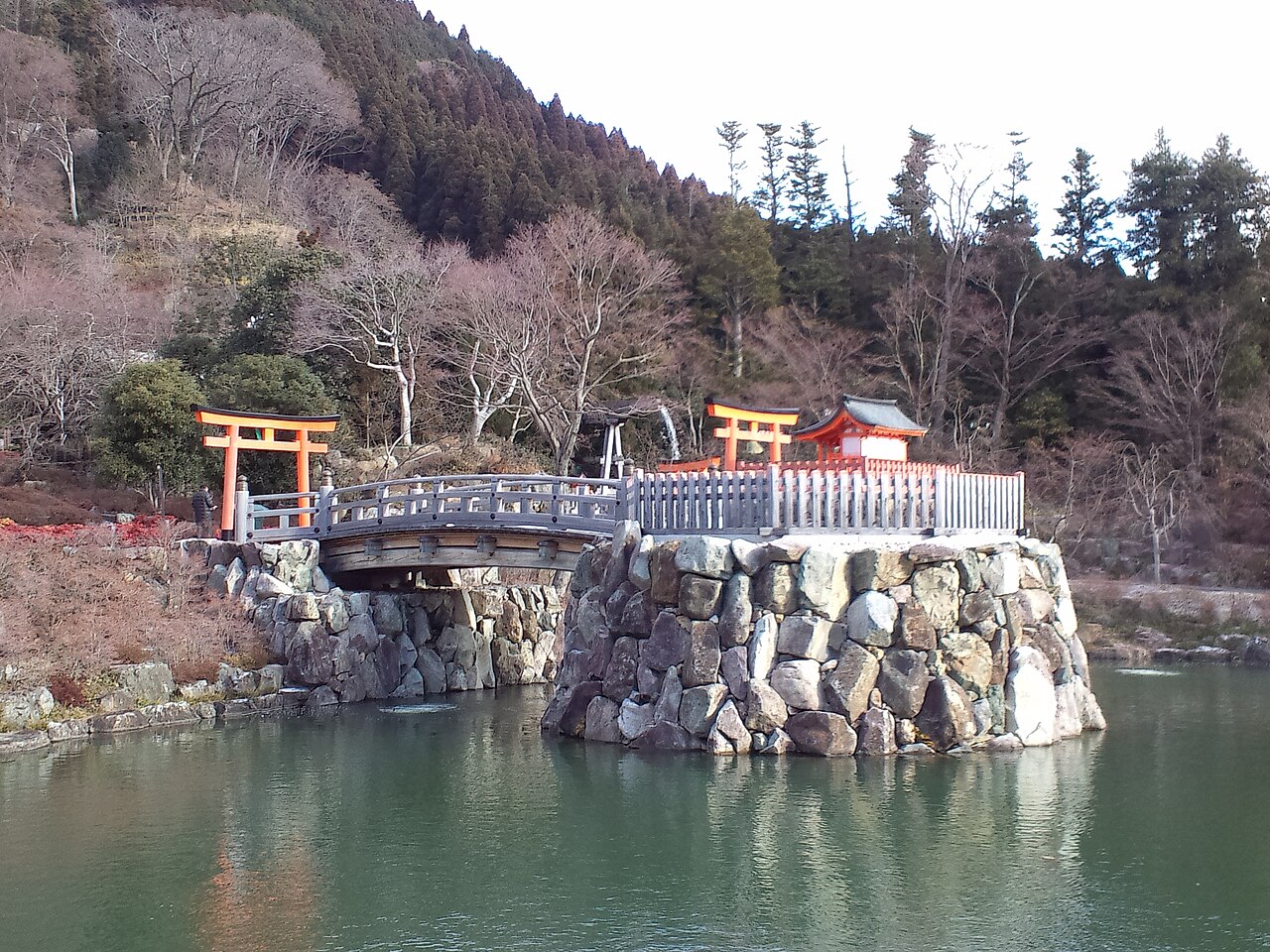 A stone-walled islet with red torii and a small shrine linked by a bridge across a pond at Katsuo-ji, with bare trees on the hillside behind