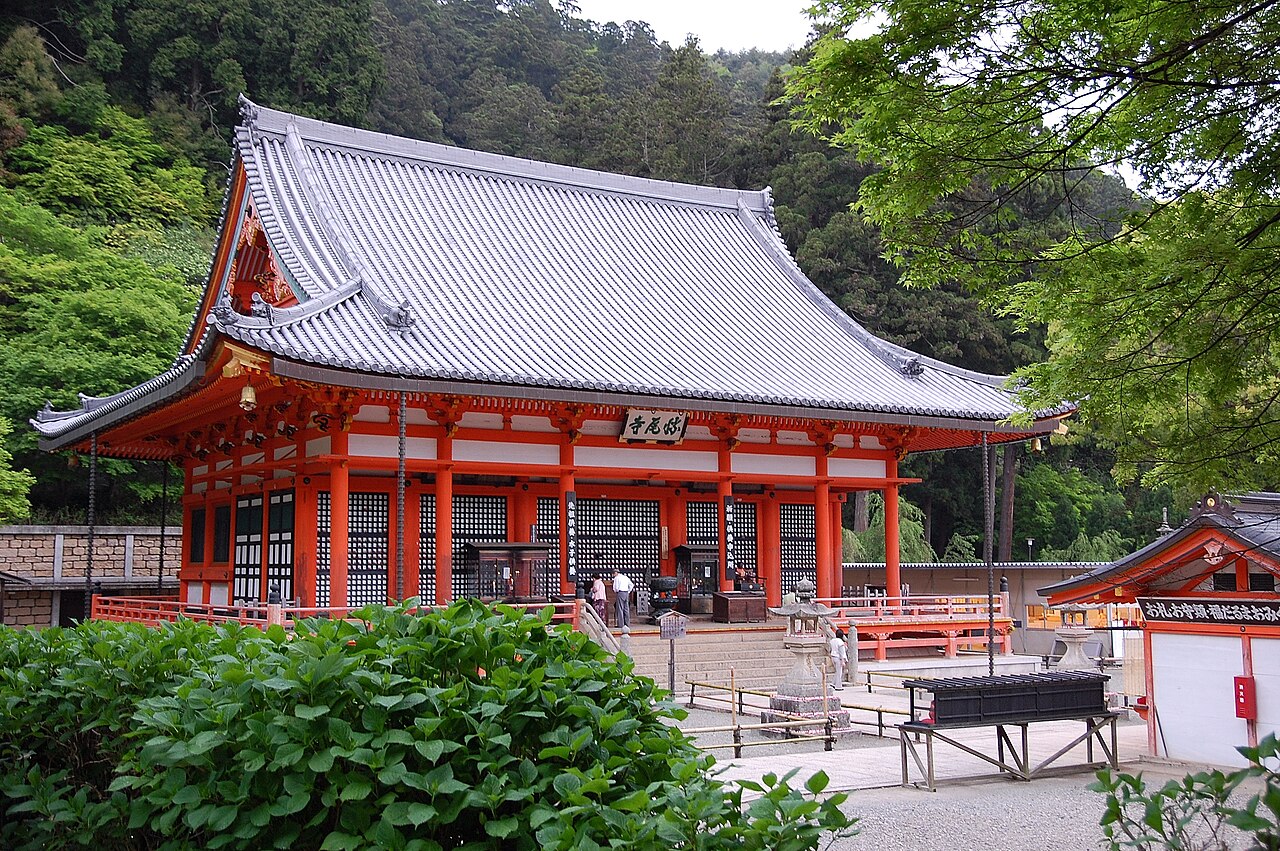 The red main hall of Katsuo-ji with a gray tiled roof, framed by green maple leaves
