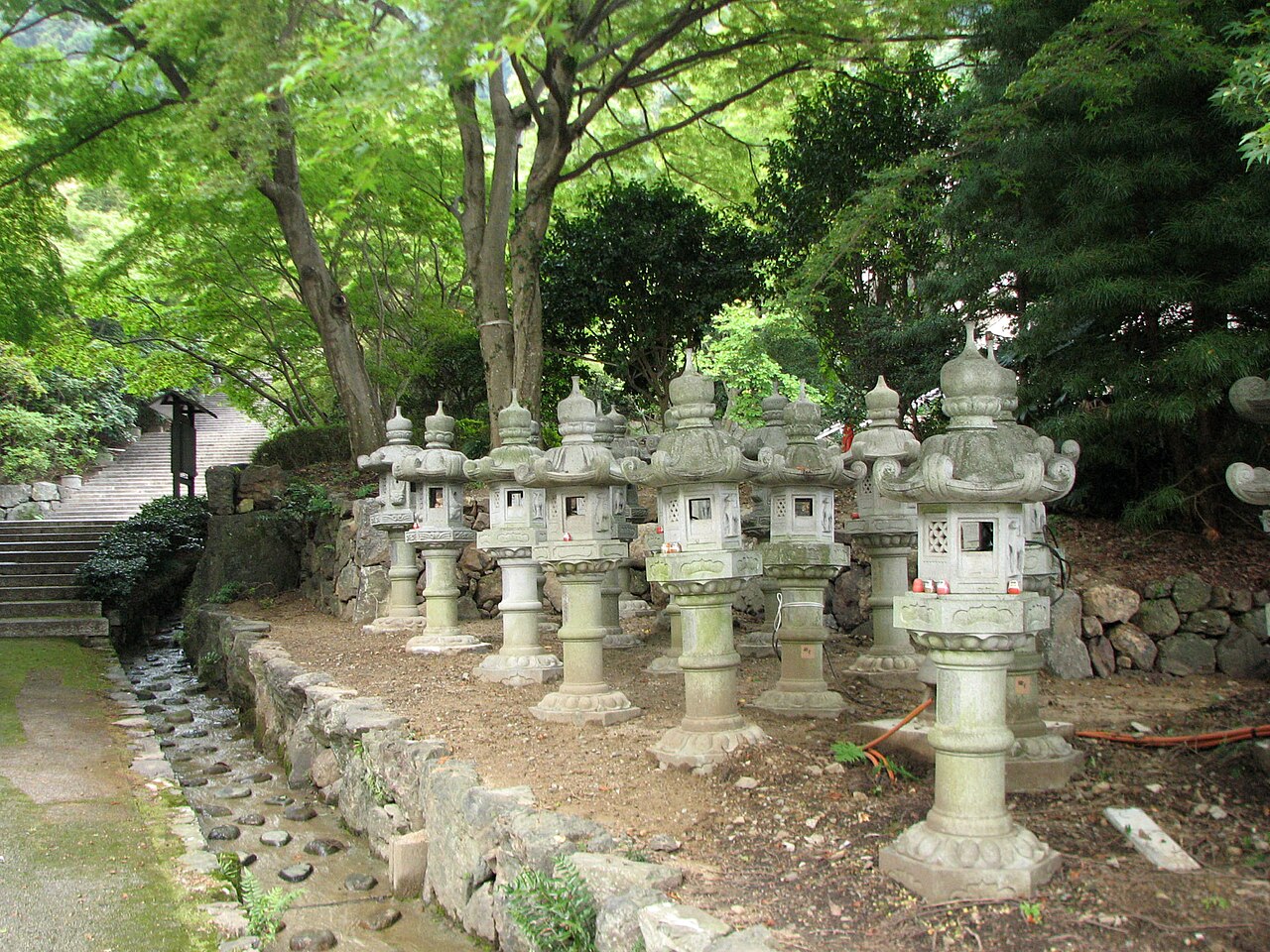 Rows of stone lanterns on a stone-edged slope among green trees at Katsuo-ji