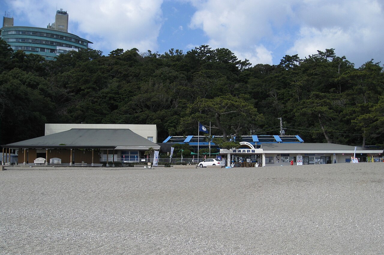 Low buildings of the Katsurahama Aquarium entrance on a gravel beach, with a pine-covered hill and a hotel tower rising behind
