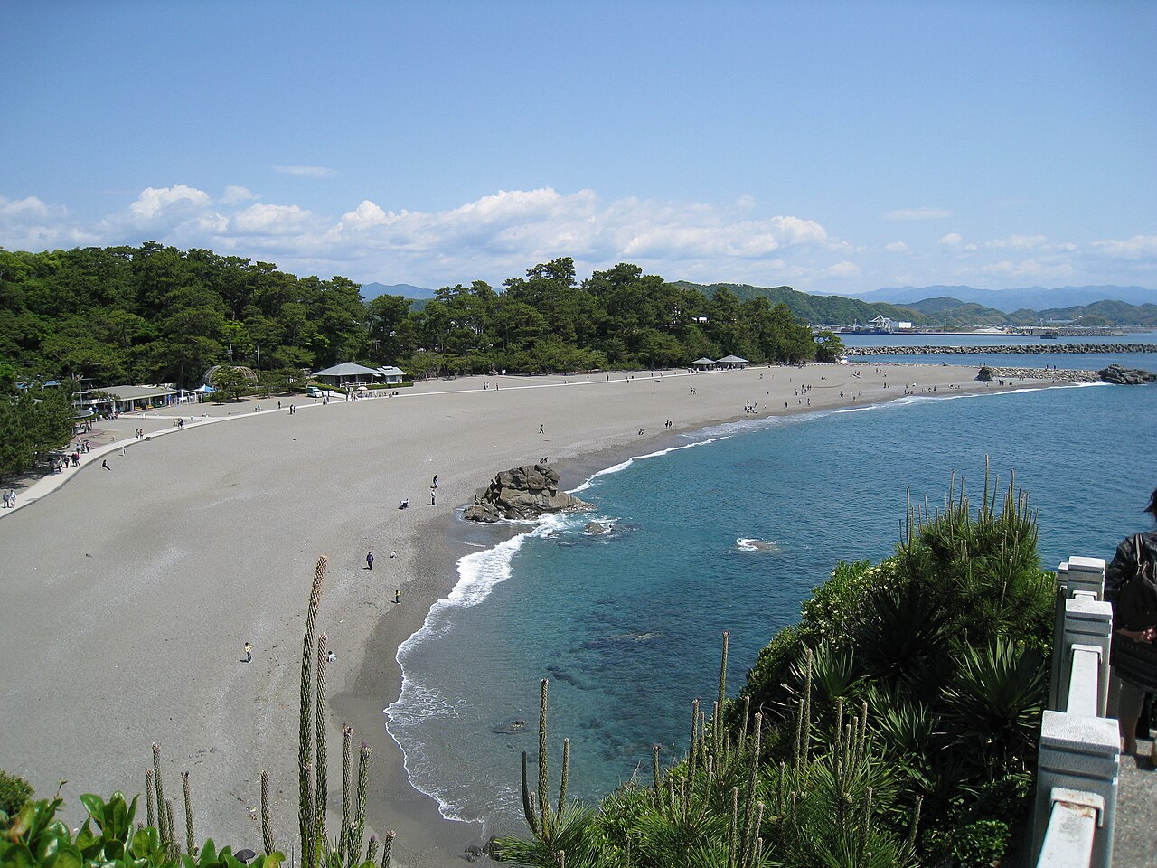 A long curved gravel beach seen from a headland, with a small rocky outcrop at the water's edge, pine woods behind and a few visitors strolling