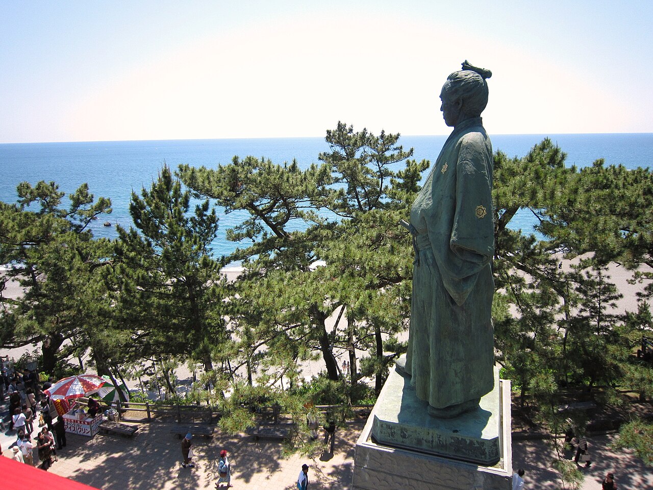 Side view of the bronze Sakamoto Ryoma statue on its stone pedestal, with pine trees below and a wide blue sea and gravel beach beyond