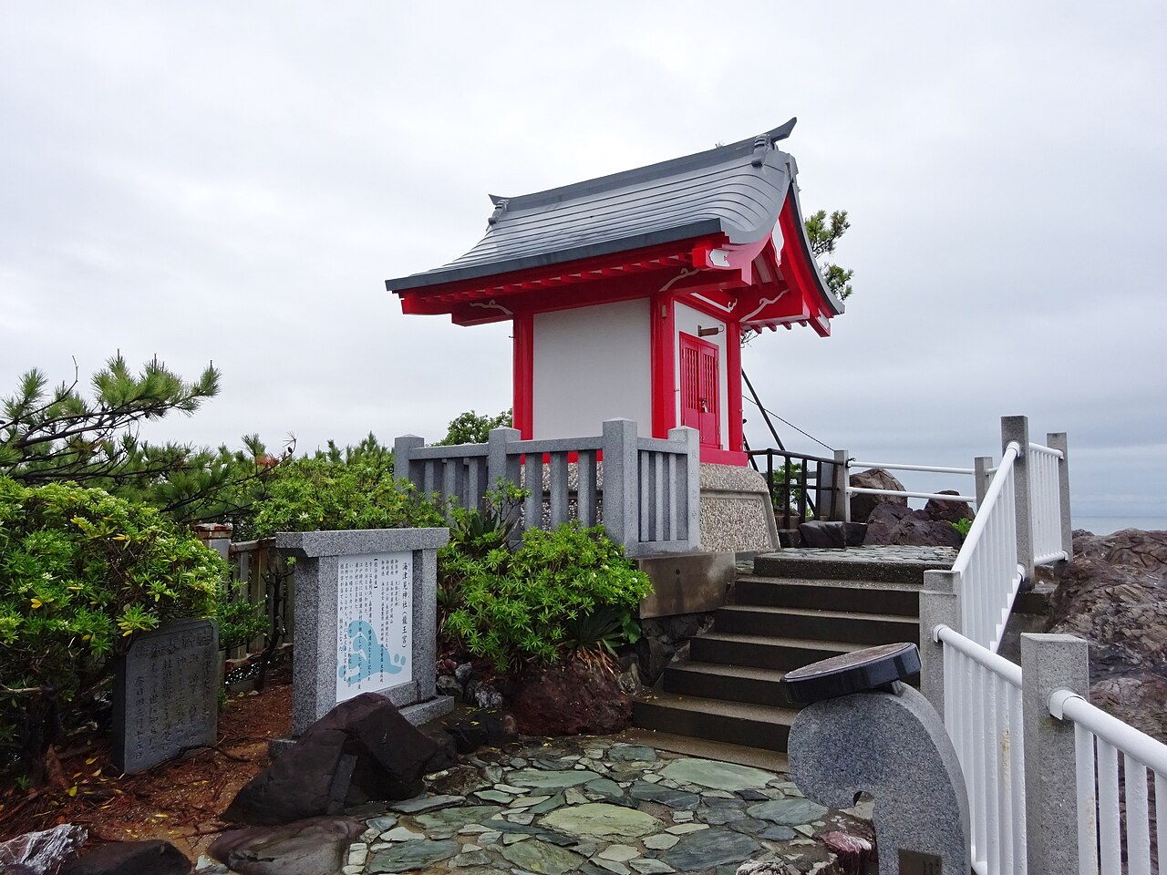 A small red-and-white shrine with a gray curved roof on a rocky headland, reached by stone steps beside white railings under an overcast sky