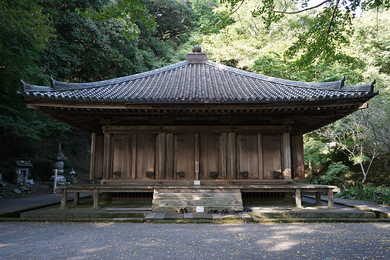 A weathered wooden hall with a low sweeping tiled roof and closed plank doors, set in a quiet wooded hollow with a few pairs of shoes left on the veranda steps