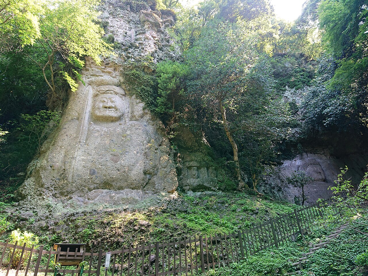 Two huge Buddhist figures carved into a mossy rock face among green trees, the larger one at left with a fierce face and a second relief tucked into an alcove at right behind a wooden fence