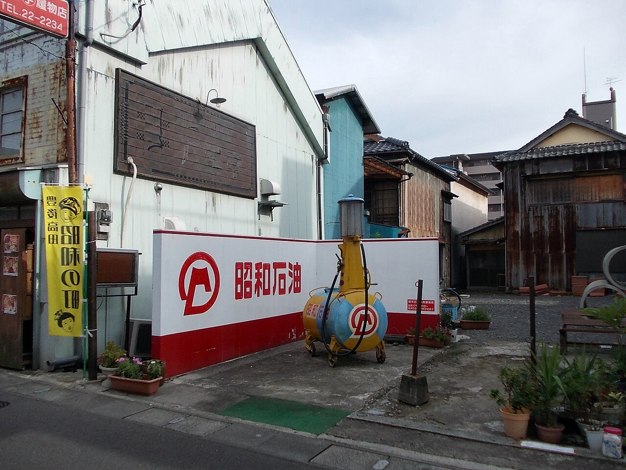A back street of old shopfronts with a large white and red wall sign, an old yellow roadside fuel pump and a yellow banner with the town's name hanging from a pole