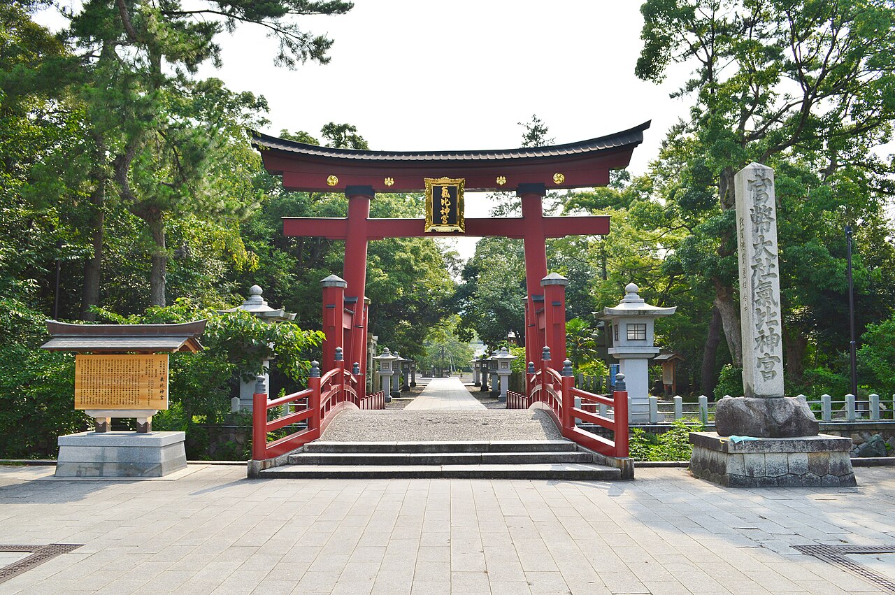 The tall vermilion torii gate of Kehi Jingu shrine standing at the end of a stone-paved approach lined with trees and stone lanterns.