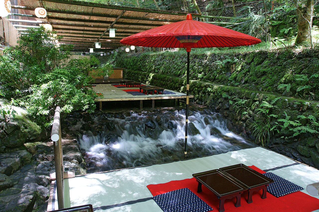 A riverside dining platform over a small stream, with a red parasol, low tables and cushions on red cloth, and green moss-covered banks.