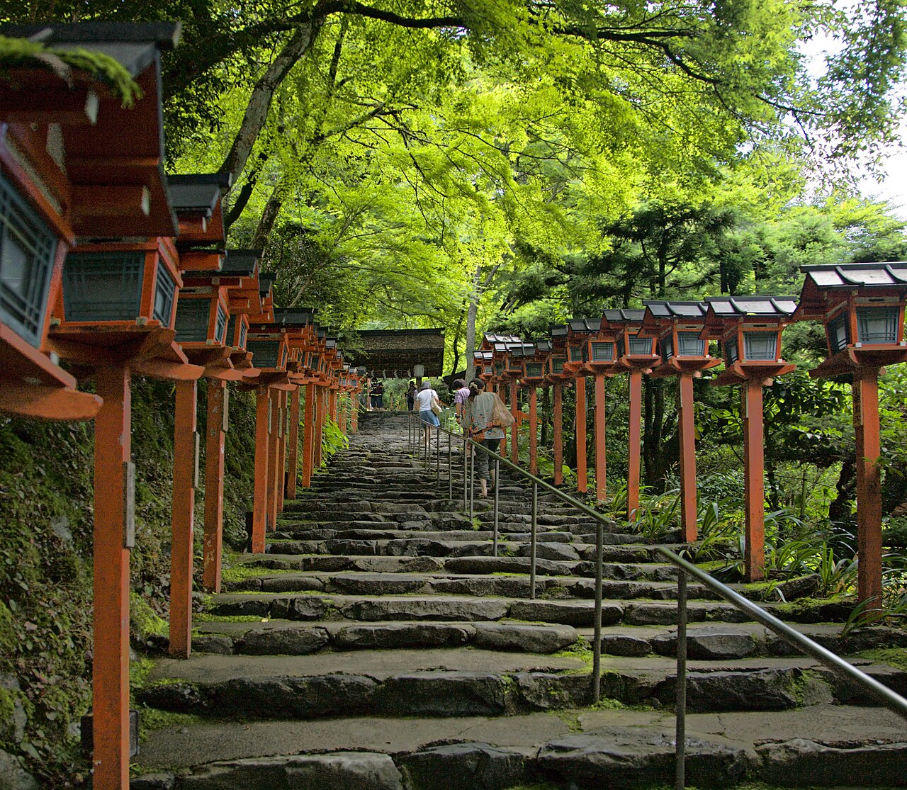 A long stone staircase lined on both sides with red lanterns, running up through bright green trees.