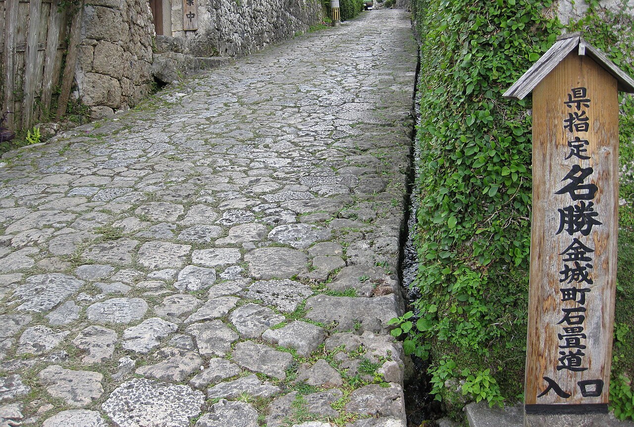 The uneven, rounded paving stones of Kinjocho stone-paved road in Shuri, Naha, bordered by a leafy vine-covered stone retaining wall, with a wooden signpost marking it as a prefecturally designated scenic spot.