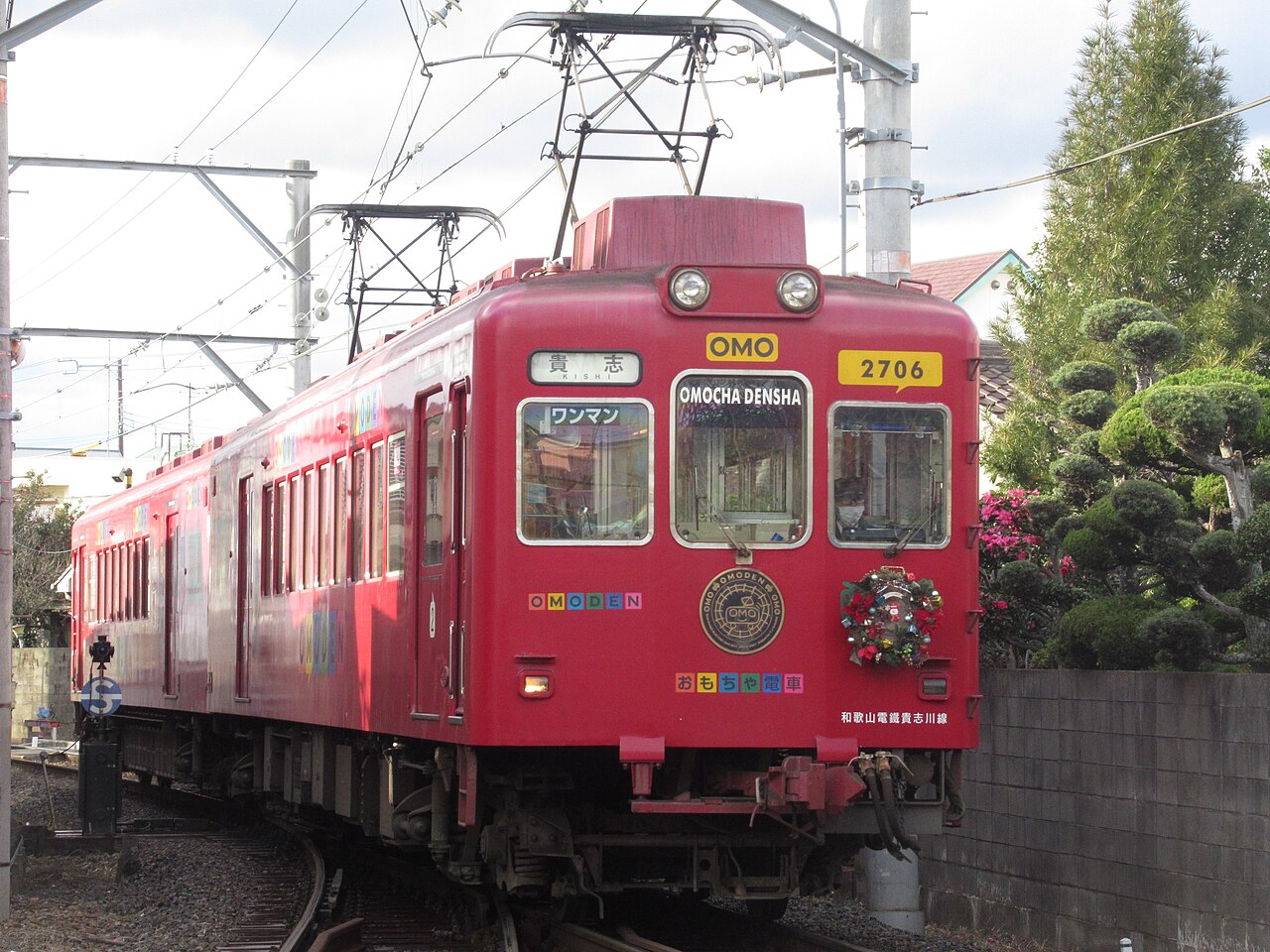 The bright red Omocha Densha toy train of the Kishigawa Line standing at a station with a small wreath on its front, trees behind it