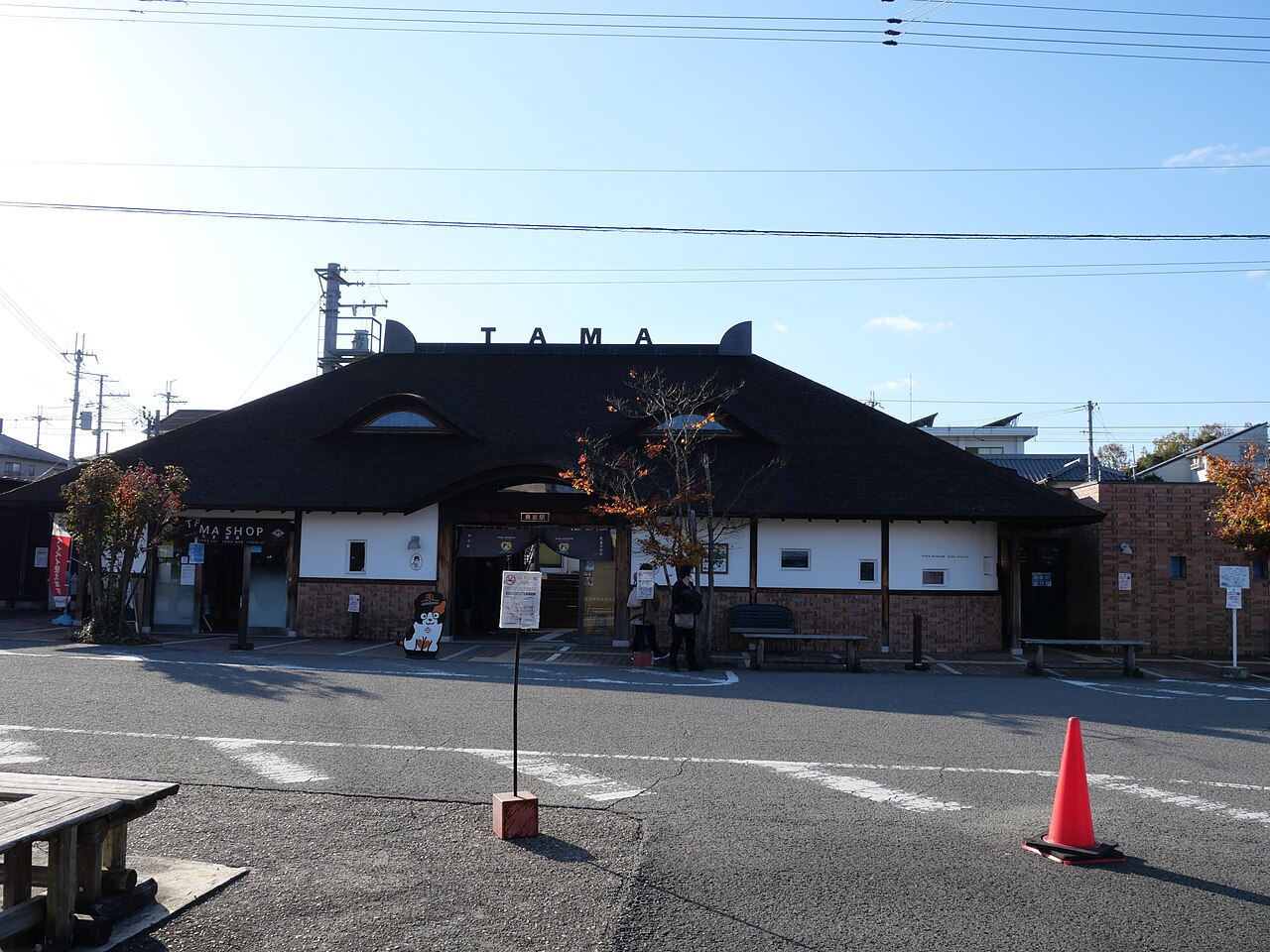 The cypress-bark-roofed building of Kishi Station with two small cat-ear gables on the ridge and the word TAMA above the entrance, seen from the forecourt under a clear blue sky