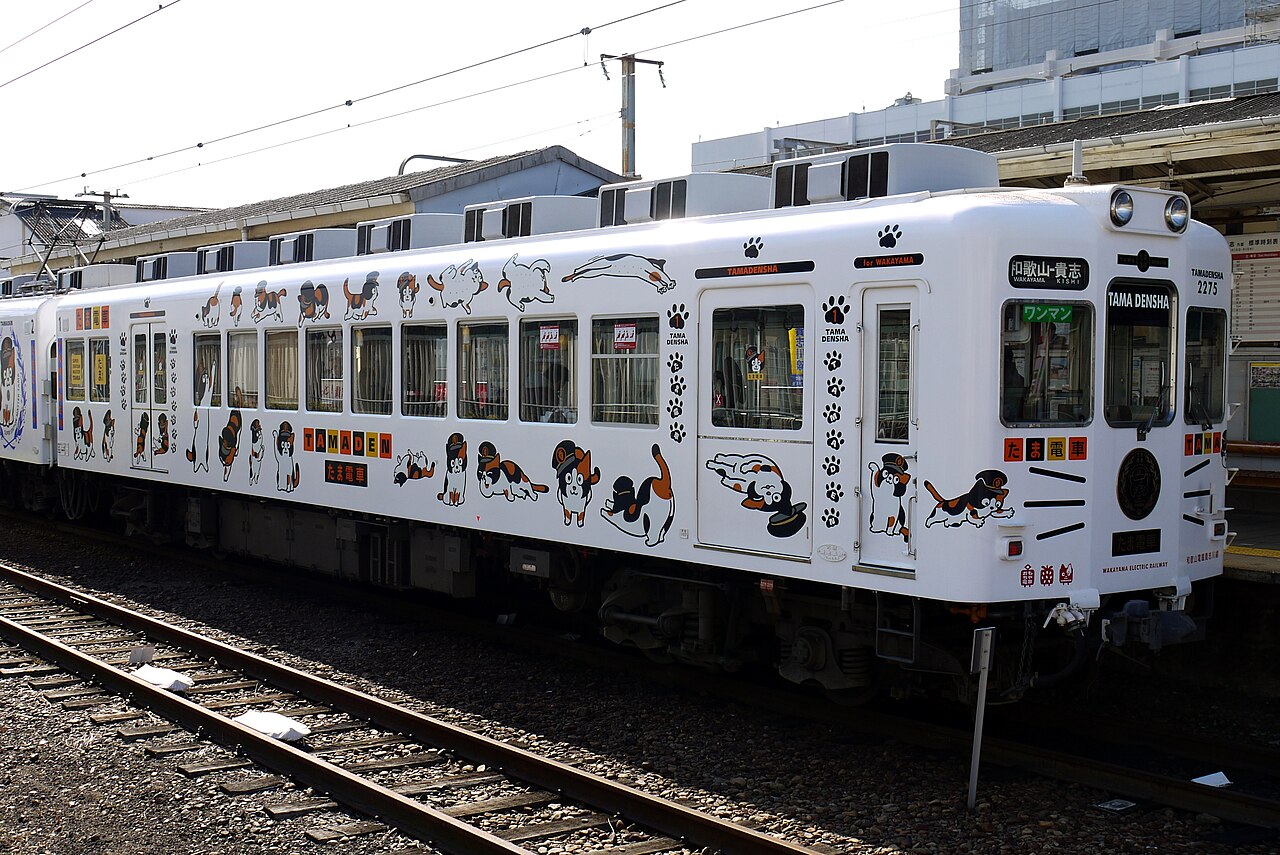 A white two-car Wakayama Electric Railway train covered in painted cats and paw prints, the Tama Densha, standing on the tracks beside a station platform