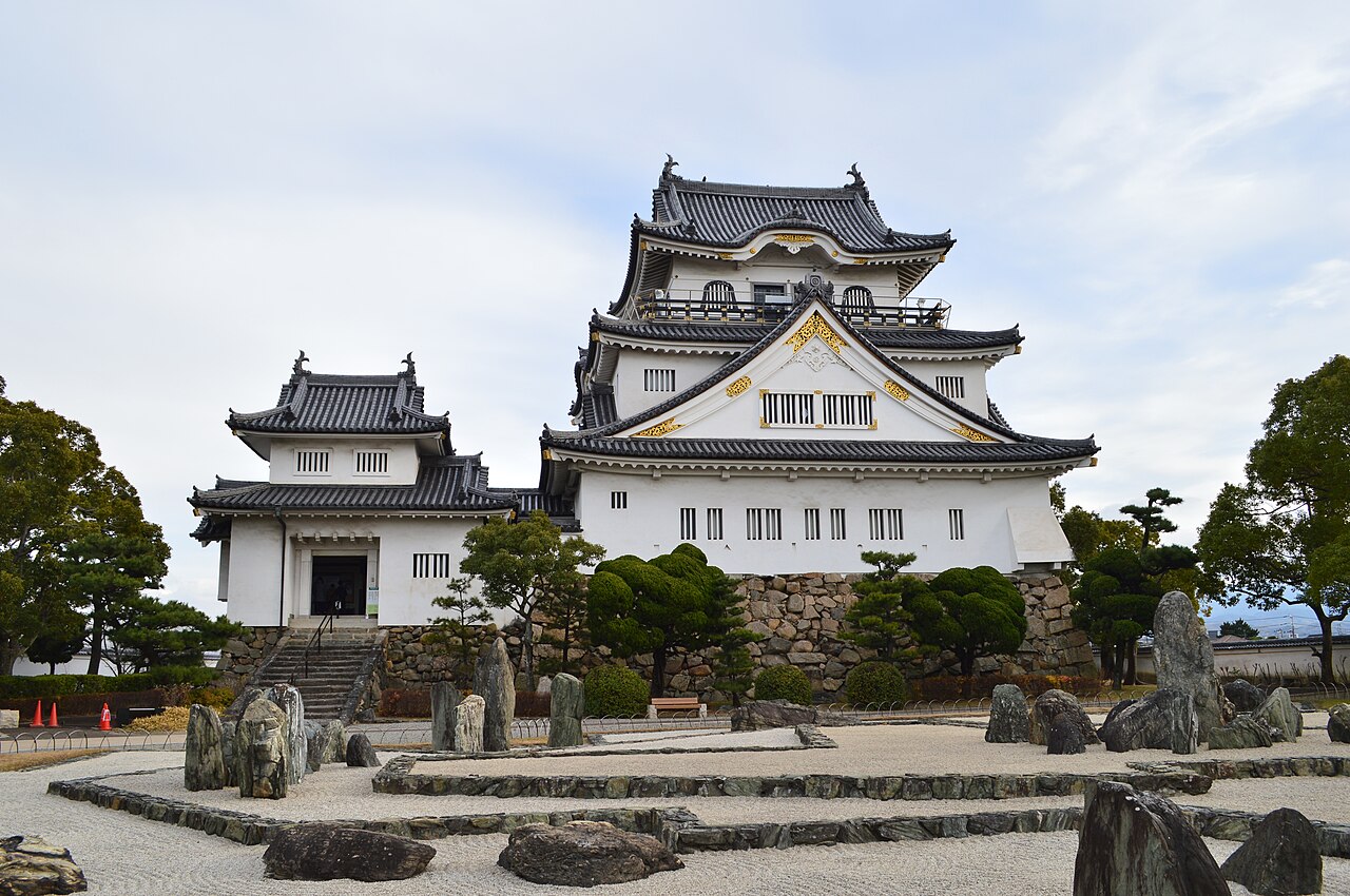 The white-walled, three-tier keep of Kishiwada Castle with gilded gables and dark tiled roofs, rising behind raked gravel and standing stones of the Hachijin no Niwa garden under a pale cloudy sky.
