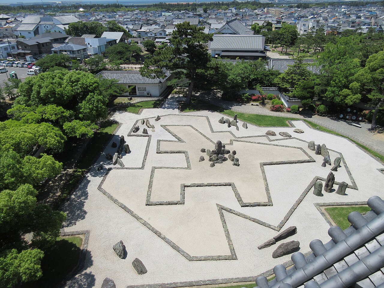 Looking down from the castle keep onto the Hachijin no Niwa garden: a large pattern of raked white gravel bordered by low stone lines, with groups of upright stones, pine trees and town rooftops beyond.
