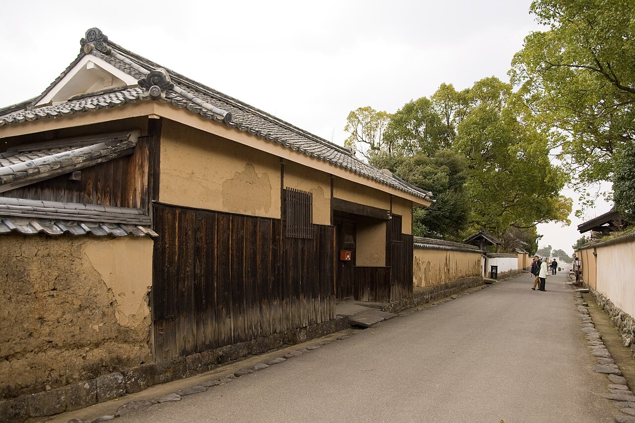 A quiet lane in Kitsuki lined with a tan earthen wall and a tiled-roof wooden storehouse, trailing off into the distance