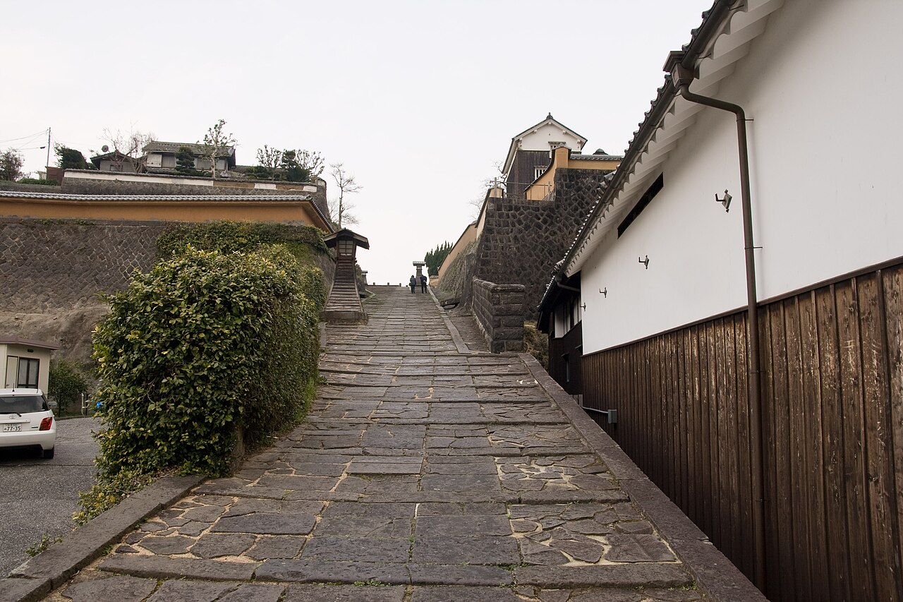 A flagstone slope rising between a white plastered wall and a green hedge in Kitsuki's old samurai quarter on an overcast day