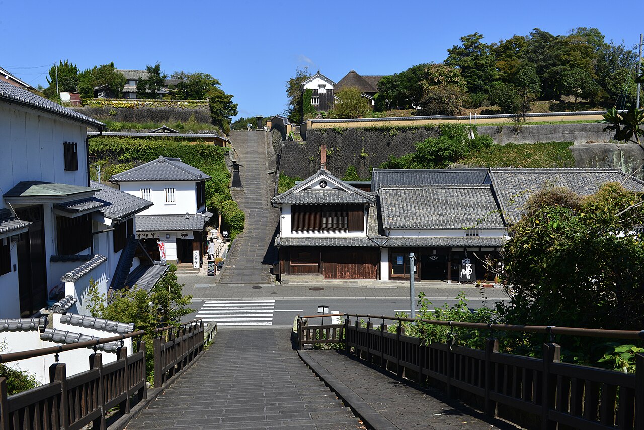 Looking across a wooden railing at the stone-paved Suya no Saka, a straight slope climbing between tiled-roof houses and earthen walls in Kitsuki