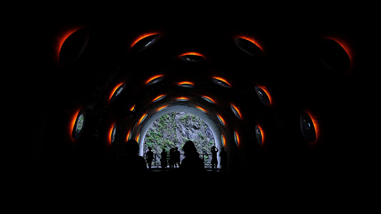 Inside the dark tunnel, rings of red light curve overhead toward a lit opening where visitors stand in silhouette.