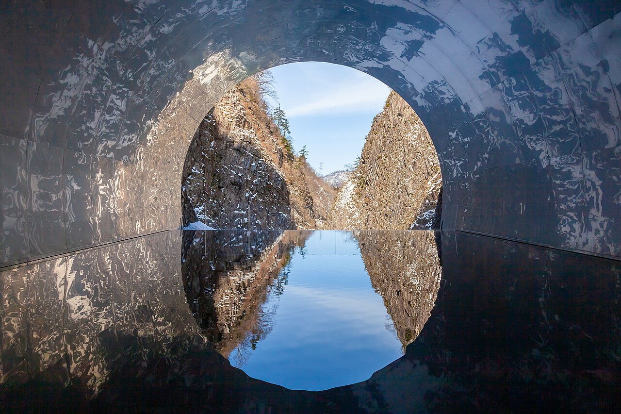The round tunnel opening at the end of the walkway frames the gorge, mirrored in a shallow pool on the floor.