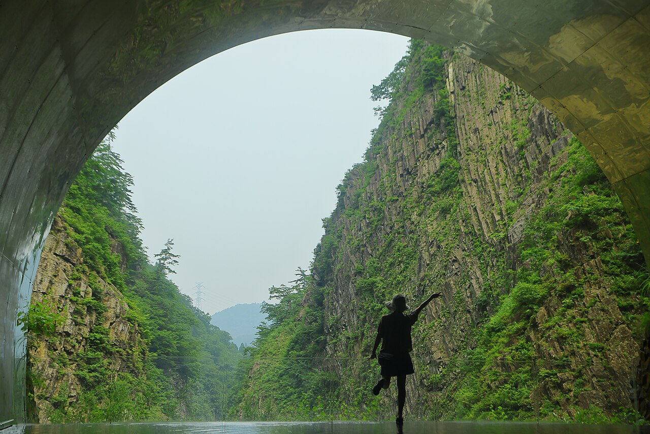 Through the arched tunnel mouth, a silhouetted figure stands before steep, green walls of columnar-jointed rock on both sides of the gorge.