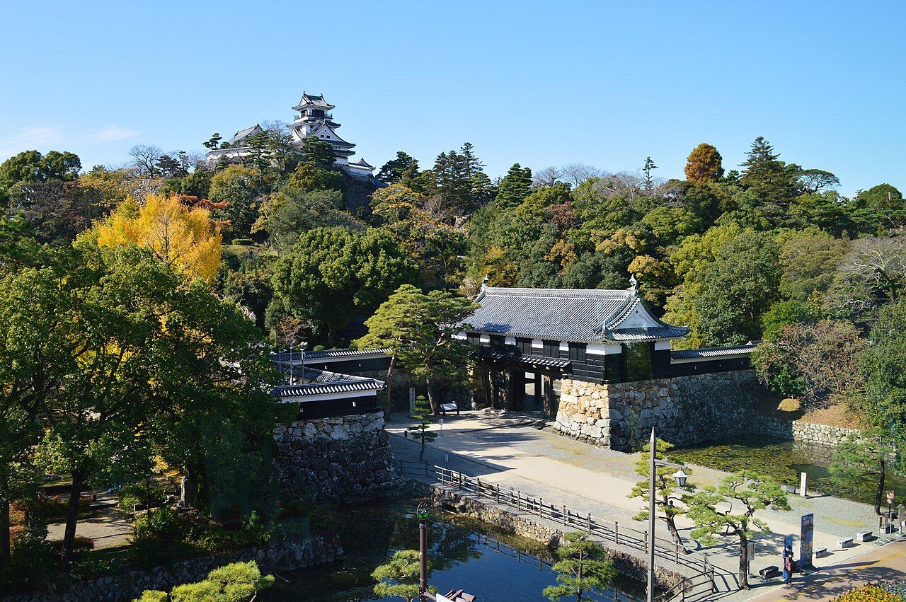 Kochi Castle keep above autumn trees, with a tiled gatehouse and stone wall beside a quiet moat in the foreground.
