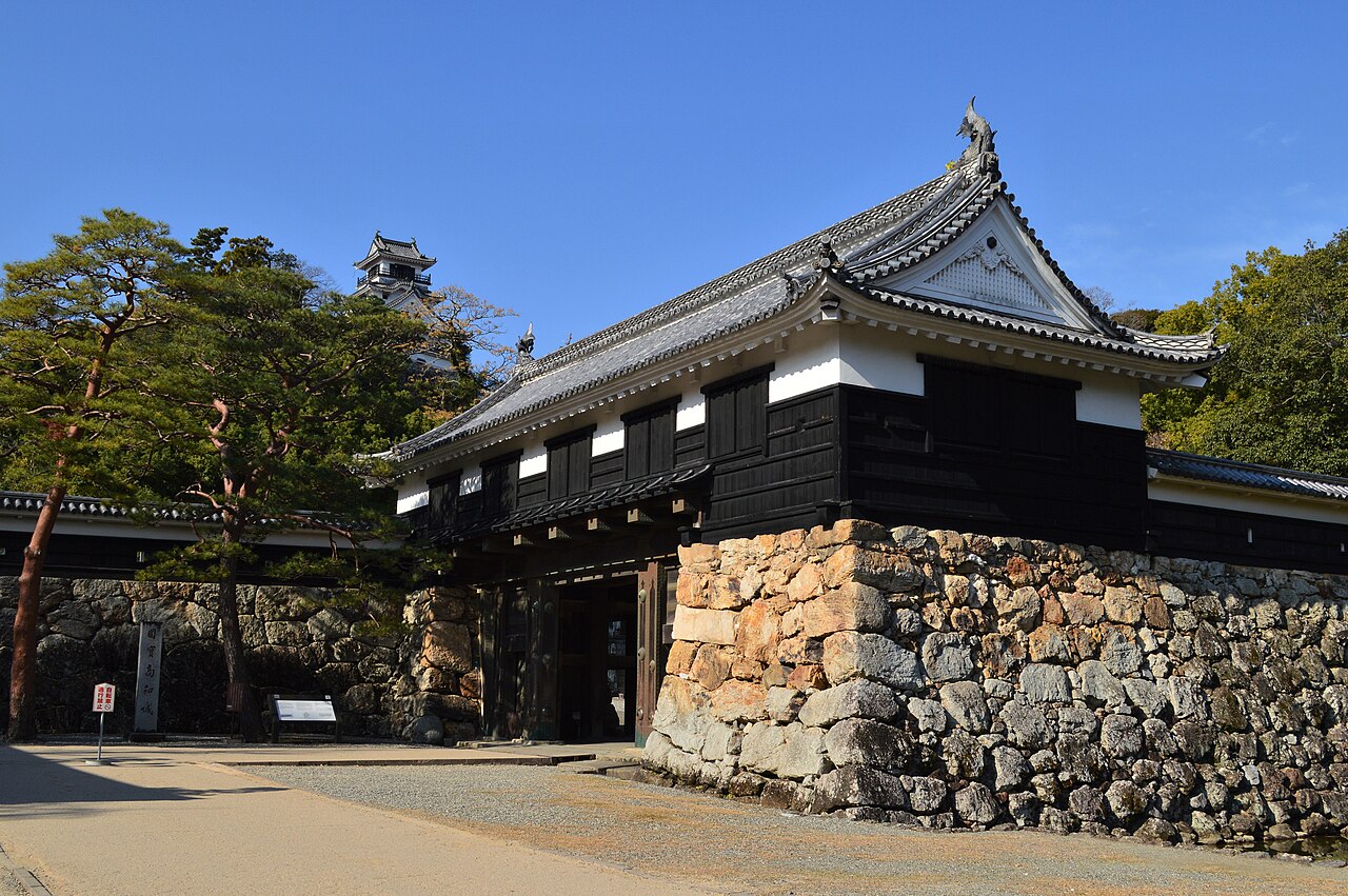 The black-and-white Otemon gate of Kochi Castle beside a rough stone wall, with the keep peeking above the pines behind.