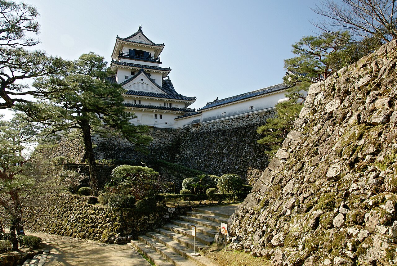 The white keep of Kochi Castle rising above a stone wall and pines, with a stone stairway leading up on a clear day.