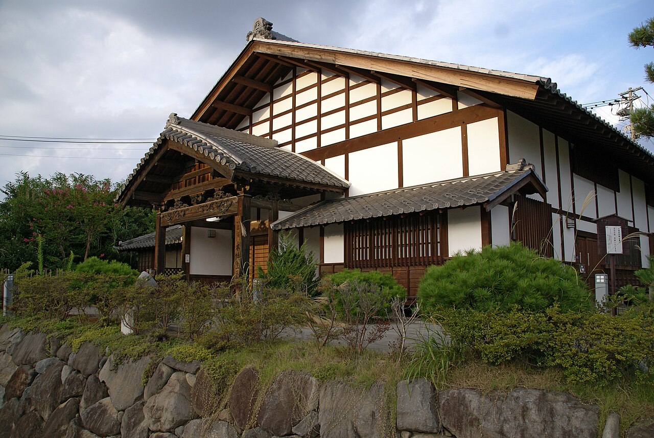 The wooden main house of the former Komoro Honjin post-station inn, with white plaster walls and a tiled roof above a stone retaining wall.