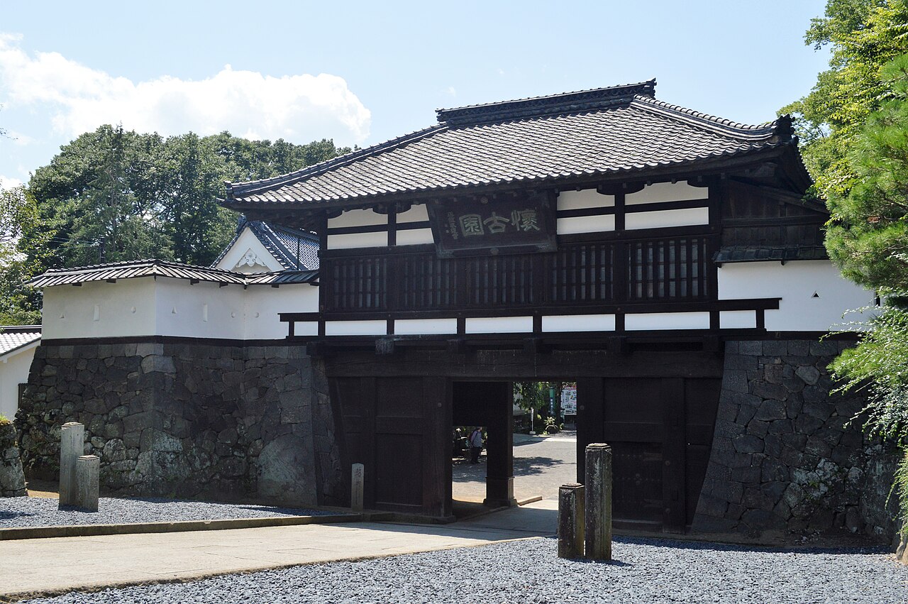 The tiled-roof Sannomon gate of Komoro Castle ruins at Kaikoen, set into a stone wall, with a plaque reading Kaiko-en above the wooden doors.