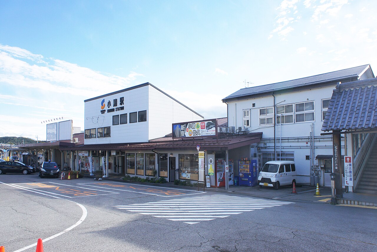 The low Komoro Station building with its name sign on the white upper wall, seen from the street across a pedestrian crossing.