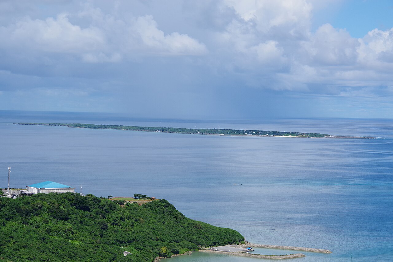 Kudaka Island lying low and flat on the horizon beyond a green headland on the Okinawa main island, under a cloudy sky.