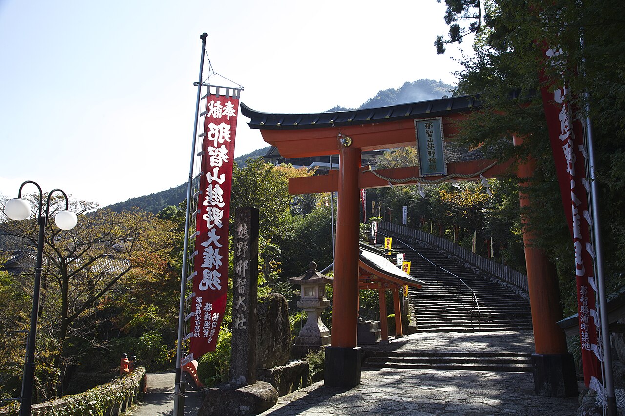 A tall vermilion torii gate bearing a nameplate for Kumano Nachi Taisha stands at the foot of a stone stairway, framed by dense green trees.