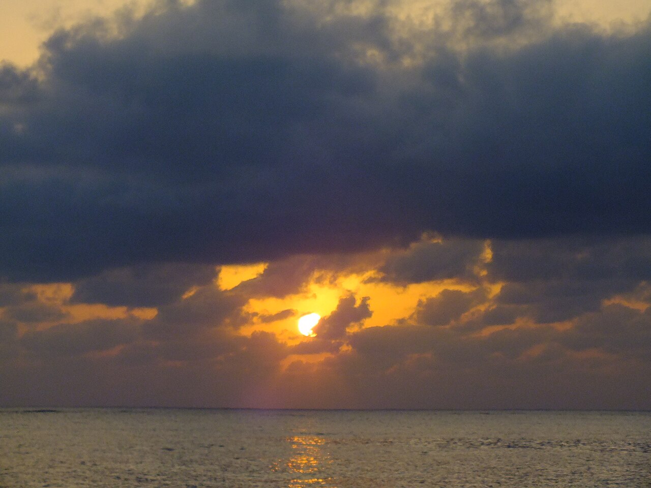 A low orange sun breaking through dark clouds over a calm sea, with a dark rocky shoreline in the foreground.