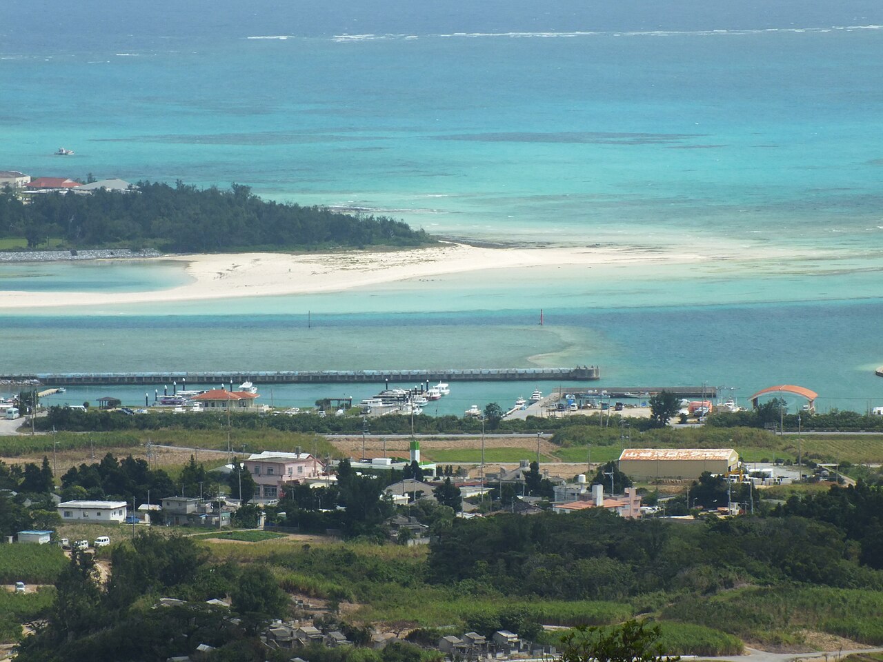 A long white sandbar curving out into shallow turquoise water off a green island coast, seen from a hill above with fields and a small harbor below.