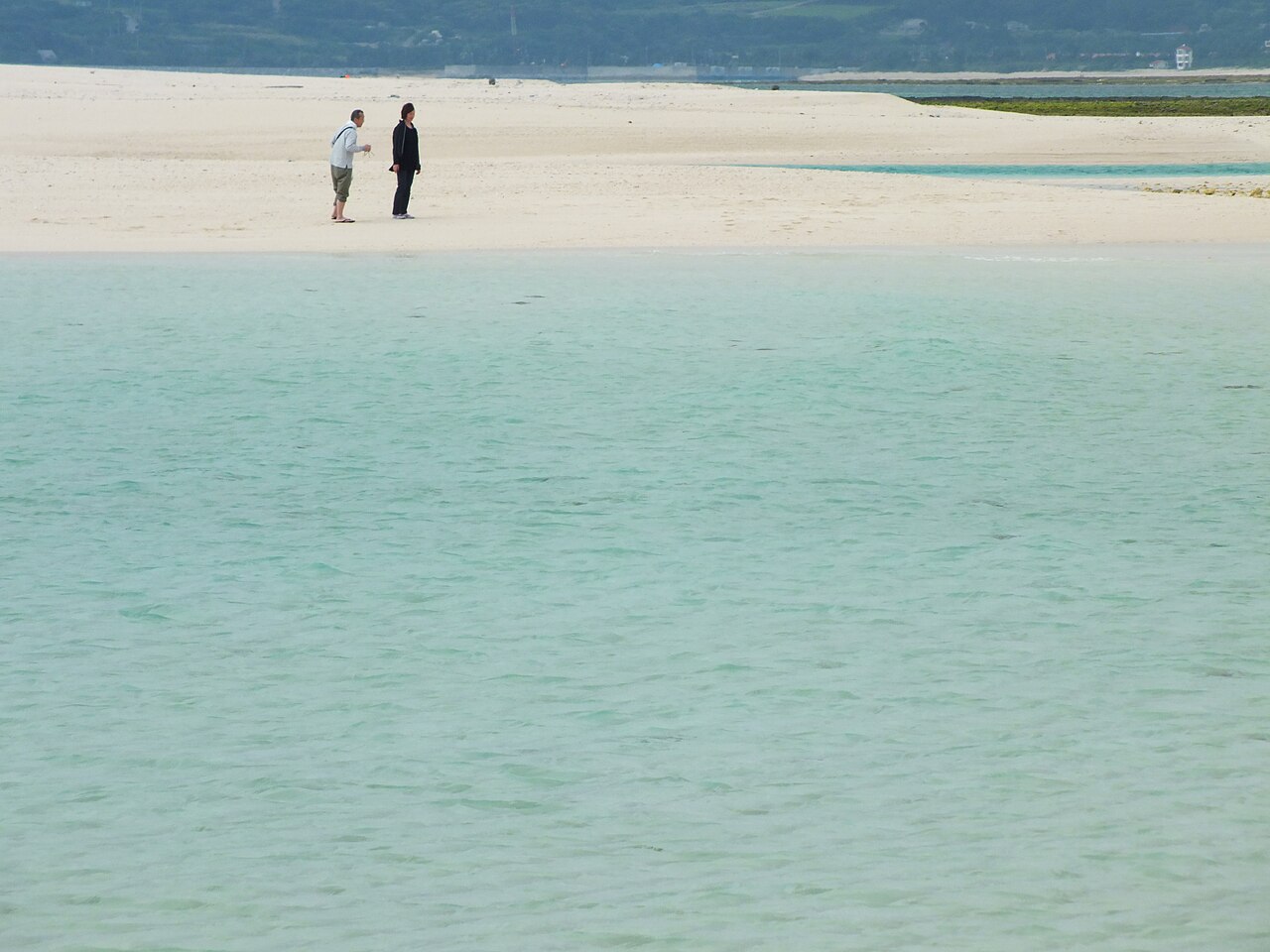 Two people walking along a pale sand spit with clear, shallow turquoise water on both sides under a hazy sky.