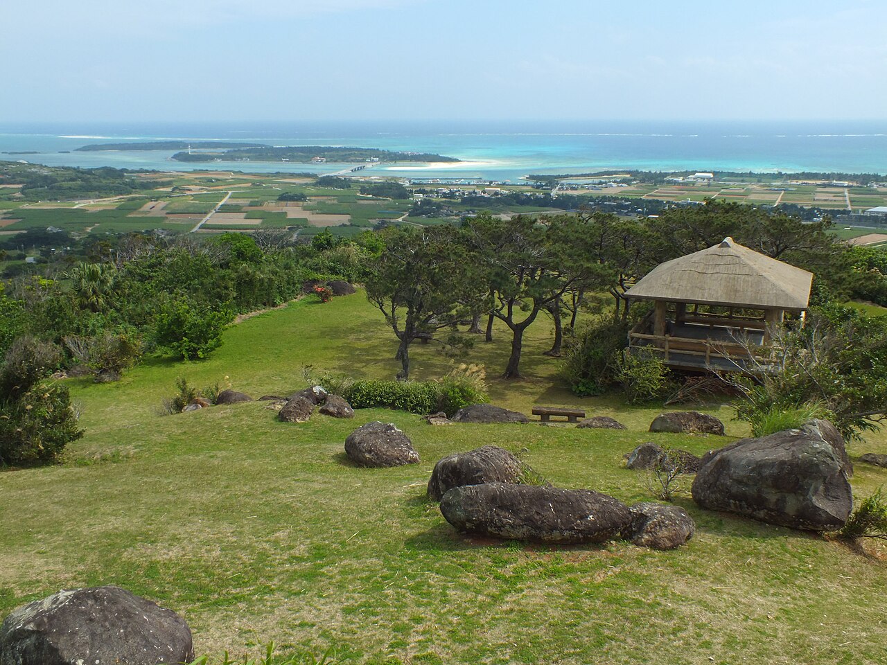 A grassy hilltop with scattered rocks and a small open-sided wooden pavilion, with the coast and sea stretching far away in the haze behind.