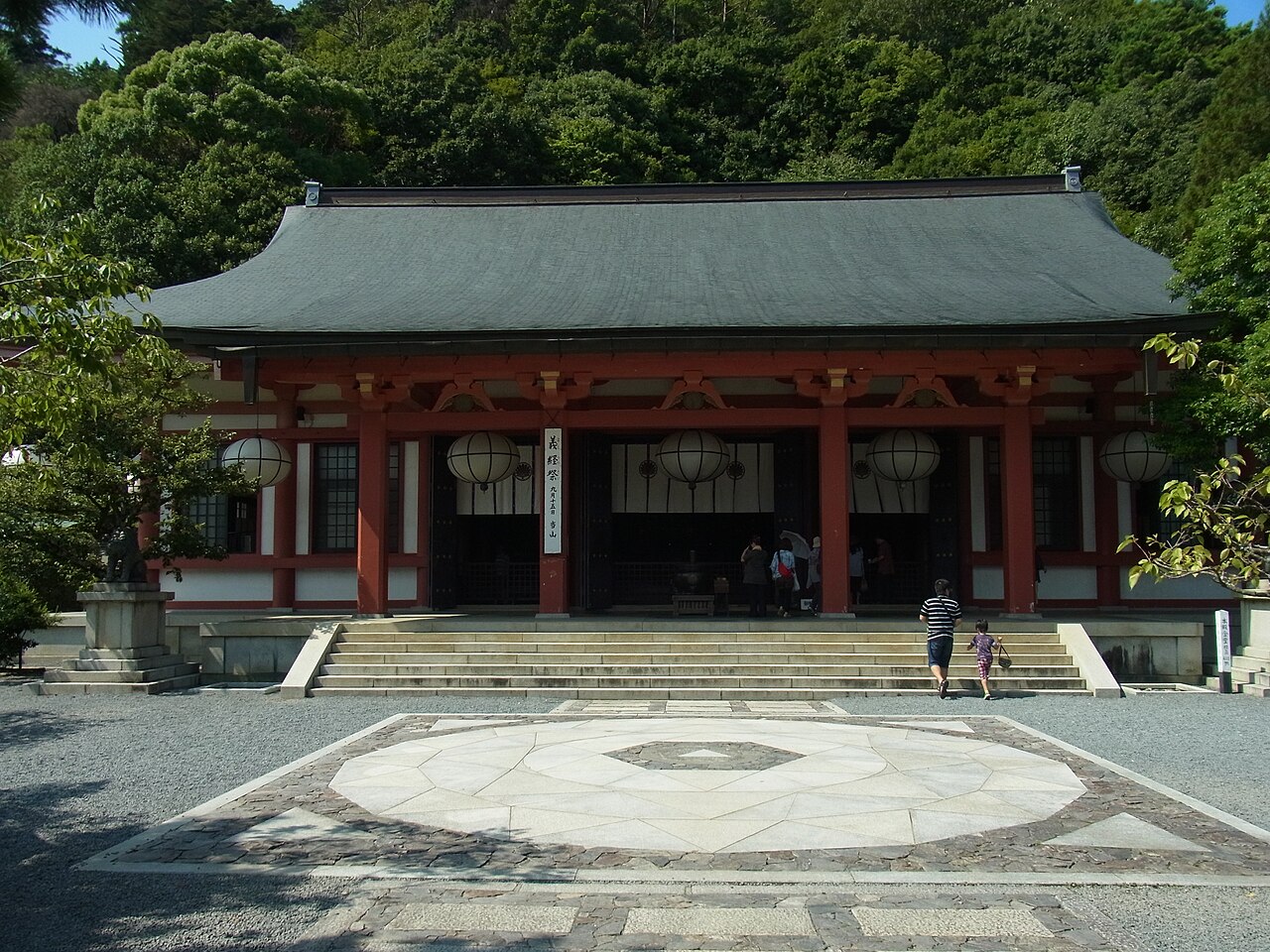 A temple main hall with a green tiled roof and red pillars, with a round patterned stone paving in the gravel courtyard in front.