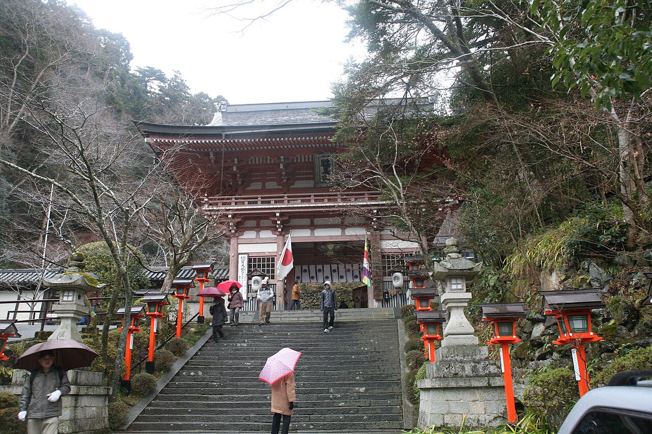 Stone steps lined with orange lanterns leading up to a red two-storey temple gate in a wooded mountainside, with visitors holding umbrellas.