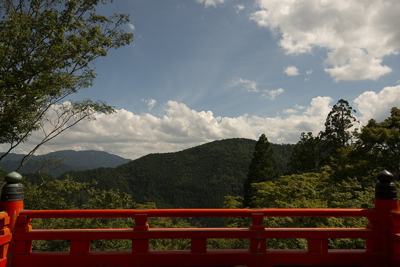 A red wooden railing with black finials in the foreground and forested mountains under a blue sky with clouds behind it.