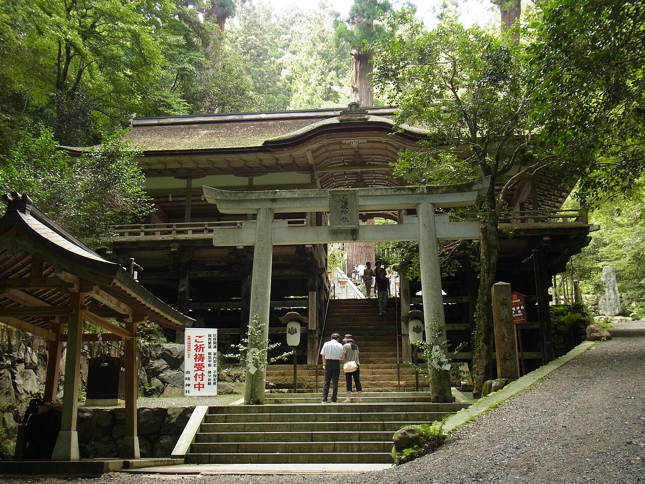A stone torii gate and steps leading to a wooden shrine hall among green trees, with a few visitors on the path.