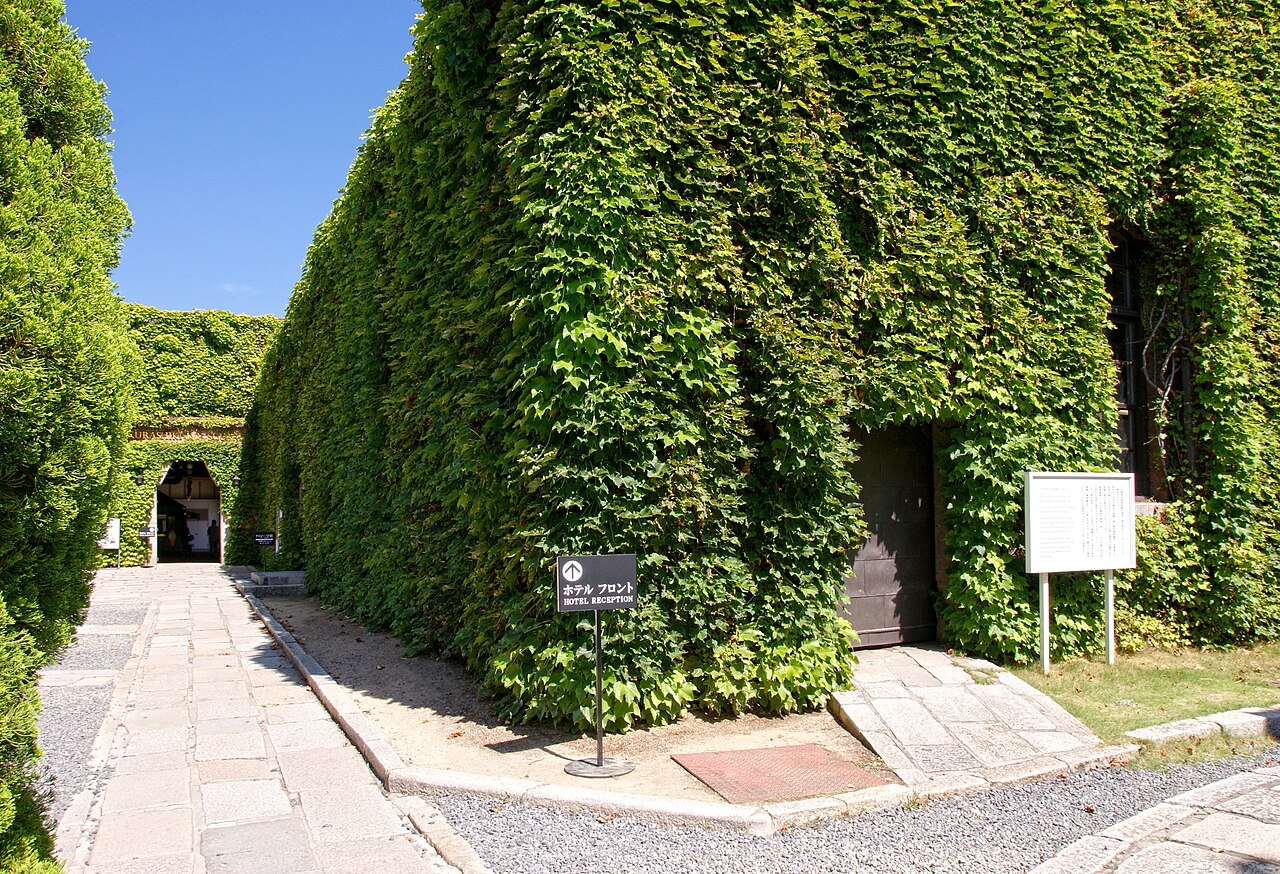 A stone-paved path beside a tall brick building wall completely covered in green ivy, with a dark passage doorway at the end.