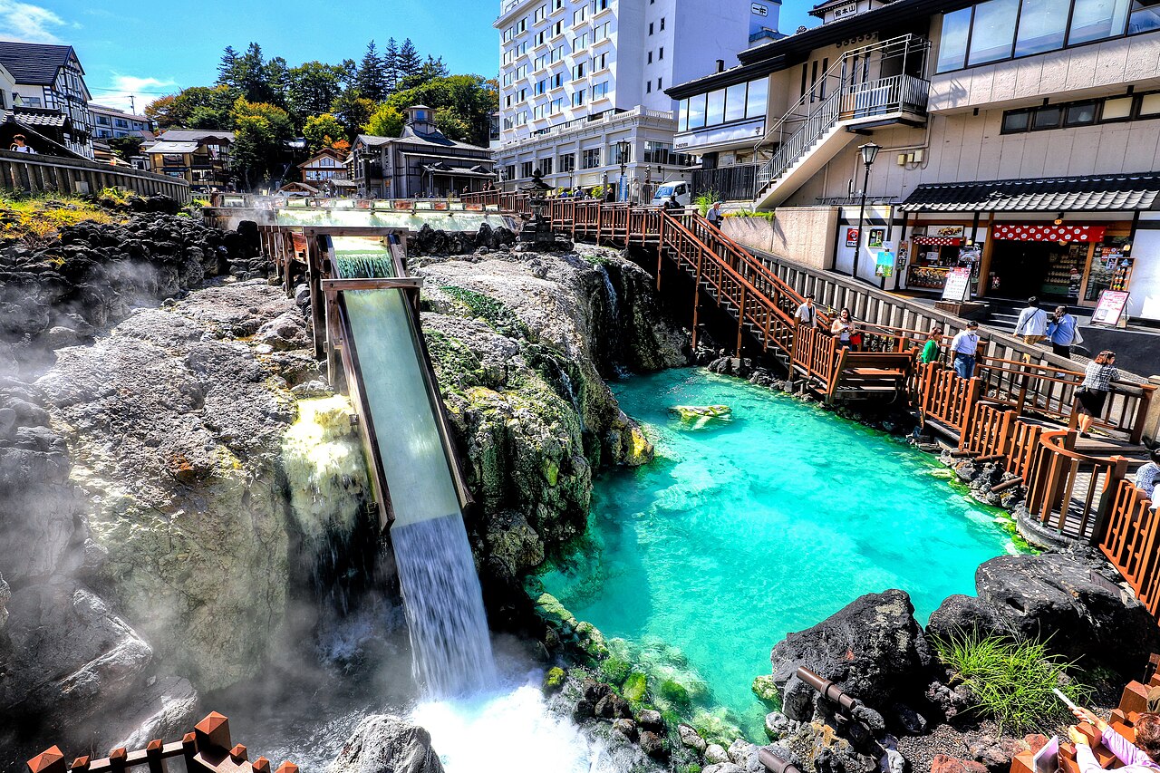 Hot spring water pouring from the end of the Yubatake flume into a turquoise pool ringed by mineral-crusted rocks, with a wooden walkway and visitors along the right.