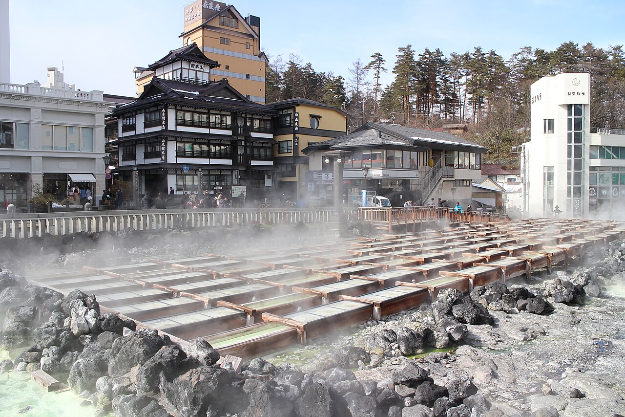 The Yubatake hot spring field in Kusatsu, rows of wooden flumes laid over dark volcanic rock with steam drifting over them and old inn buildings and a white tower behind.