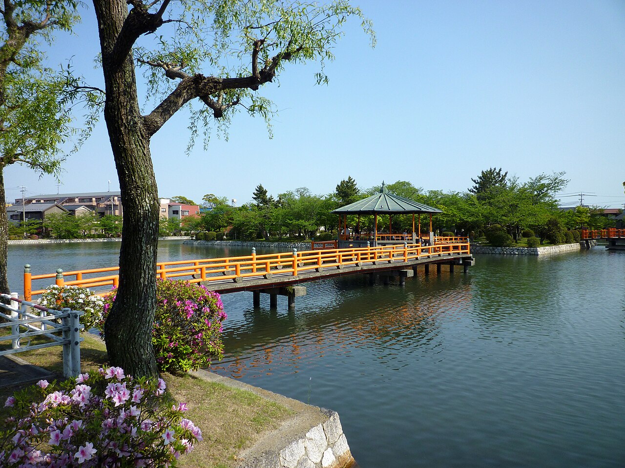 An orange-railed wooden walkway crosses a calm moat to a green-roofed hexagonal pavilion among trees, with pink azaleas and a tree trunk in the foreground and houses beyond under a clear blue sky.